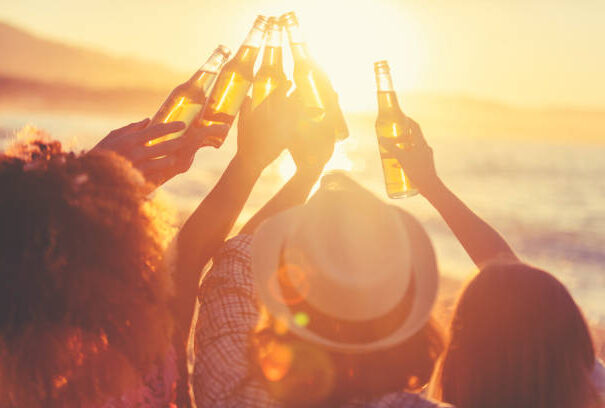 Group of young people partying on the beach at sunset. They are all holding bottles of beer and celebrating with a toast. Silhouette with the beach, sun and sea in the background.